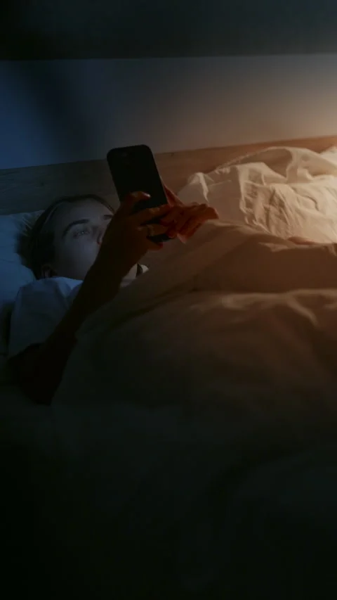 Young female using smartphone in bed at night, illuminated by soft light Stockbeeldmateriaal 341525325