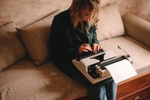 Young female writer using typewriter while sitting on sofa at home Stock Photos