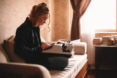 Young female writer using typewriter while sitting on sofa at home Stock Photos