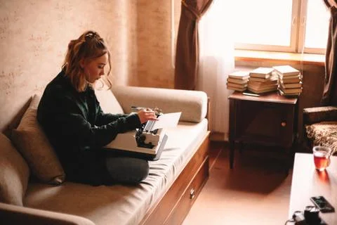 Young female writer using typewriter while sitting on sofa at home Stock Photos