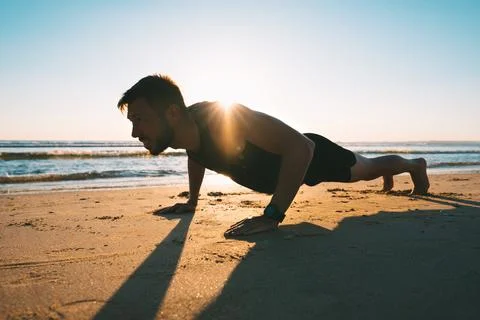 Young fit man doing push ups or exercising on the beach during sunrise Stock Photos