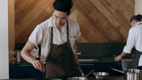 Young focused chef pouring salt in a pan, surrounded by other chefs preparing fo Stock Footage 199461855