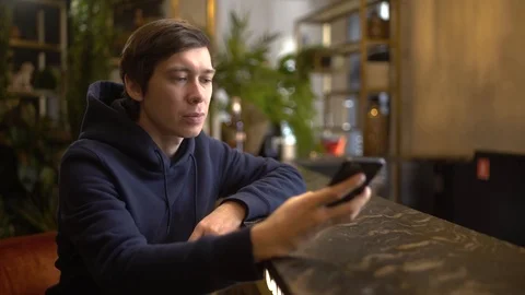 Young freelance sitting at a table in a cafe using iphone and laughing, watching Stock Footage 118964498