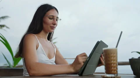 Young freelancer draws with stylus on tablet at tropical cafe, palm trees. SMM Stock Footage 252070099