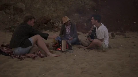 Young friends sitting by the fire on the beach in the evening, grilling sausages Stock Footage