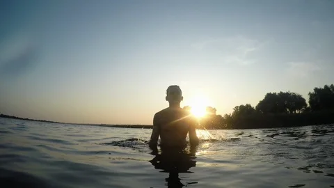 Young funny happy guy standing in the river's water and splashing it at sunset 库存影片 114667940
