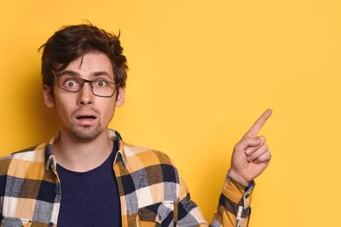 Young funny nerd guy pointing to free copy space for text on yellow studio wall Stock Photos