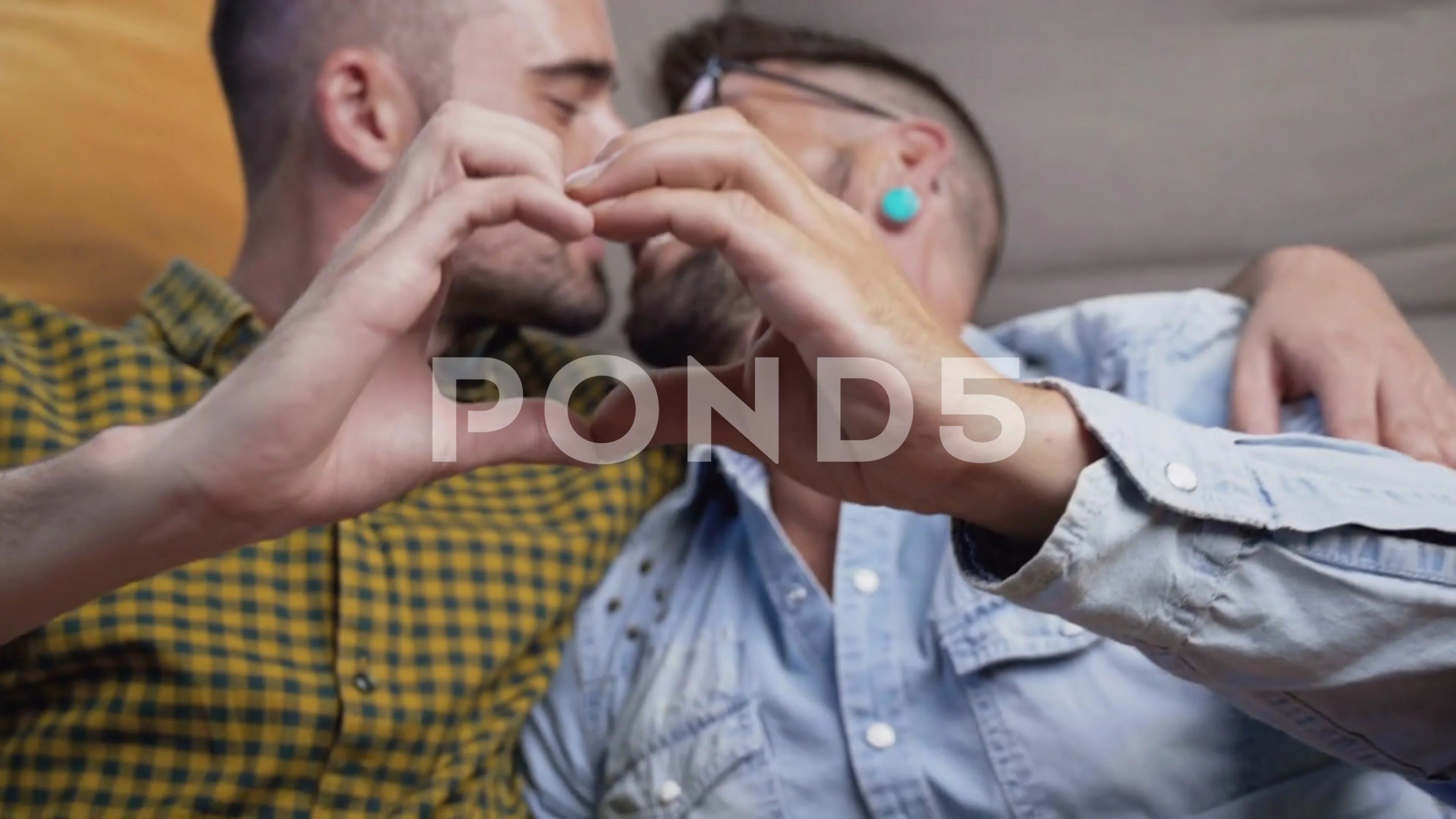 Young gay couple kissing while doing heart symbol with hands at home