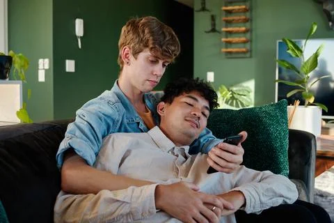 Young gay couple using mobile phone while embracing on sofa at home Stock Photos