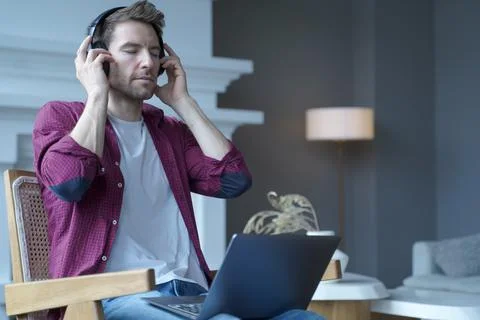 Young german guy sitting on wicker chair with laptop on his lap listening music Stock Photos