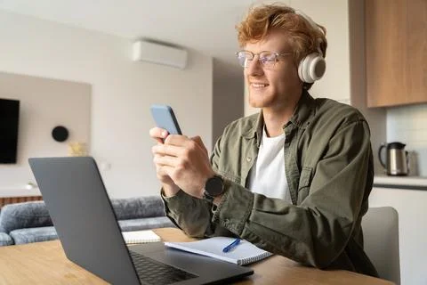 Young ginger man using smartphone while working online on laptop at home office Stock Photos