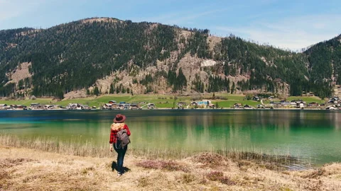 Young girl with backpack and in hat comes to the shore of mountain lake. Stock Footage 107398932