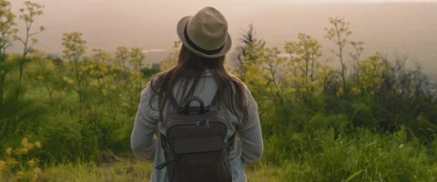 A young girl with backpack and a hat walking through the field at sunset Stock Footage 127097856