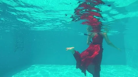 Young girl with braids in long red dress swims under water in swimming pool Stock Footage