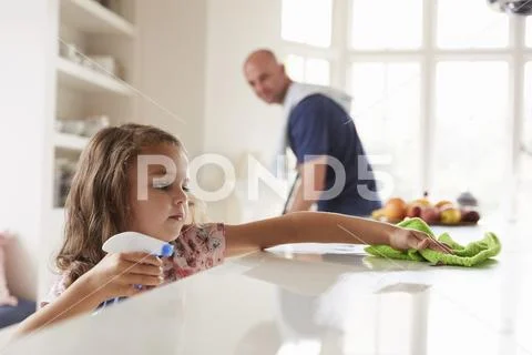 Young girl cleaning kitchen worktop, dad in background Stock Photos