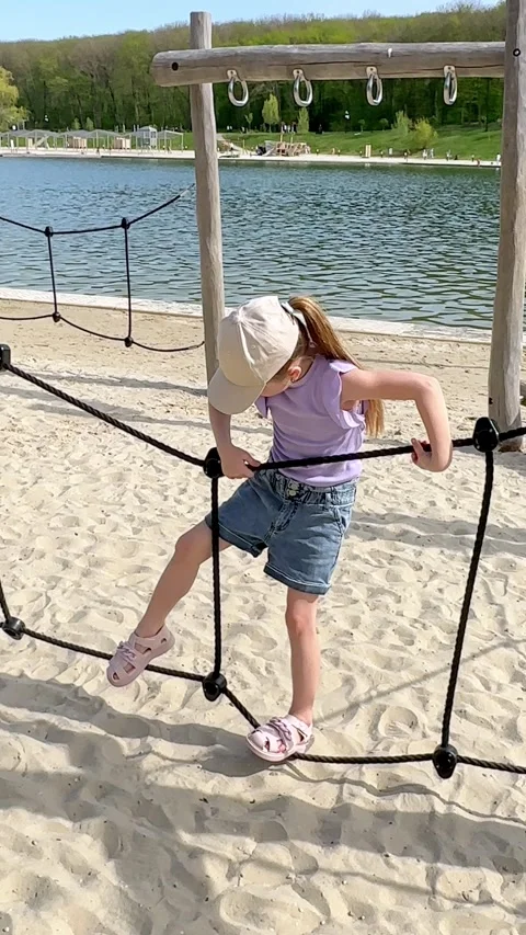 Young girl climbs on a playground structure by a lake, with a sandy beach Stock Footage 340653638
