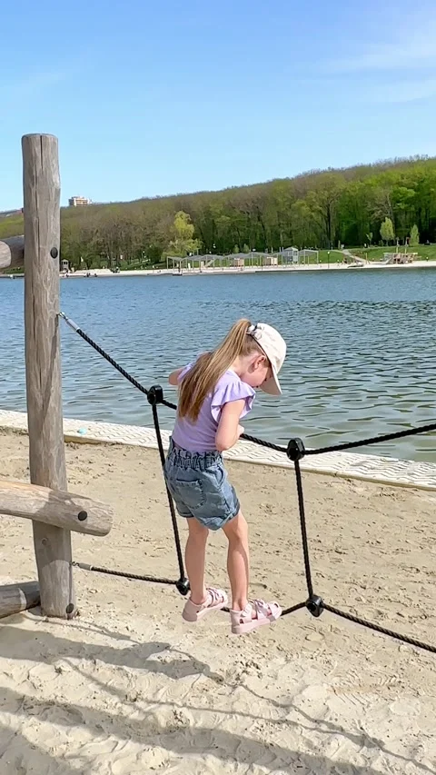 Young girl climbs on a playground structure by a lake, with a sandy beach Stock Footage 340653958