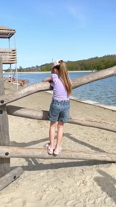 Young girl climbs on a playground structure by a lake, with a sandy beach Stock Footage 340654565