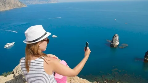 A young girl does selfie next to the blue sea sitting on a mountain Stock Footage