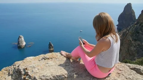 A young girl does selfie next to the blue sea sitting on a mountain Stock Footage 80659942