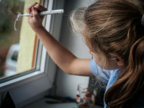 A young girl draws on a window using a white marker. Stock Photos