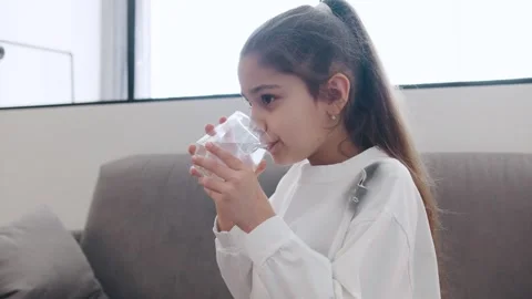 Young girl drinks water while sitting on a couch in a bright room with large win Stock Footage 330773295