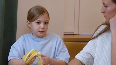 Young girl explaining something to her mother while peeling a banana Stock Footage 317847971