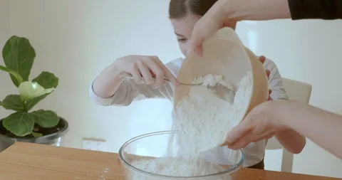 Young girl making bread dough laughs happily as her mother pours flour 库存影片 114801337
