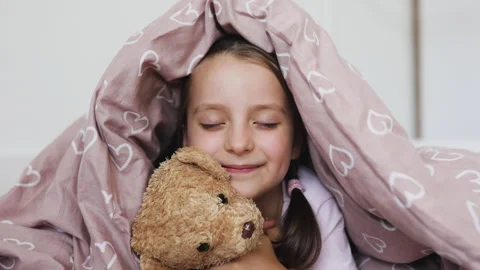 Young girl with missing teeth cuddles teddy bear under cozy blanket Video stock 295256845