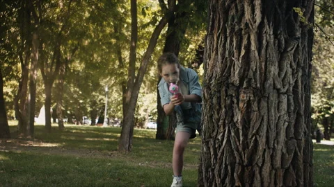 Young girl plays with water gun in park hiding behind tree during summer day Vídeo Stock 344569953