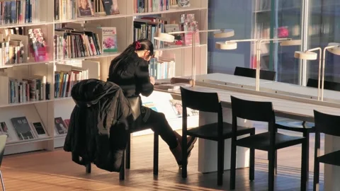 Young girl reads a book while sitting in a city library. Bookshelves in the 스톡 동영상 264125048