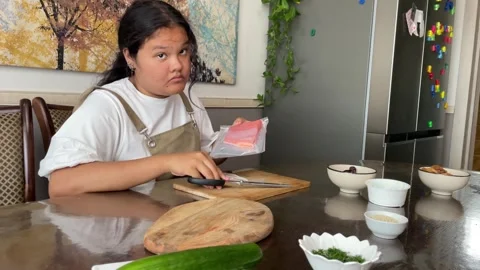 Young girl is seen using scissors to open a package in kitchen, illustrating the Stock Footage 279923272