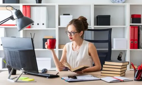 A young girl sits at a computer table and holds an open book and a red cup in Stock Photos