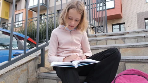 Young girl sits on steps while reading textbook 스톡 동영상 284663309