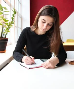 A young girl sits at a table in a cafe and writes something in a notebook.  Stock Photos