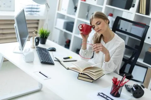 A young girl sits at a table in the office and holds a cup in one hand, and in Stock Photos