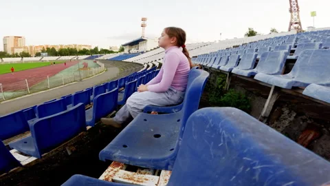 Young girl sitting alone in an empty stadium watches a sports game unfold in Stock Footage 290457178