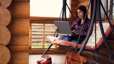 Young  girl sitting on a swing and works remotely on laptop in a country house Stock Footage