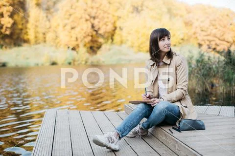 A young girl sketching near a lake in the autumn forest. Stock Photos