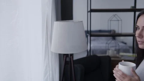 A young girl is standing by the window drinking coffee and looking into the 스톡 동영상 129330601
