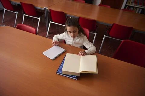 A young girl studies at a library table, with an open book and notebooks, cap Stock Photos