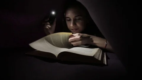 Young girl using flashlight in dark for reading book under the blanket Stock Footage