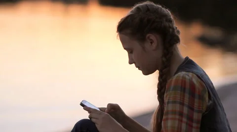 Young girl using smartphone reading news on social media by the lake at sunset Stock Footage 65798008