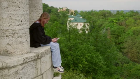Young girl using smartphone sitting on ancient ruins overlooking green forest Stock Footage 295282832