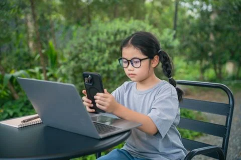 Young girl using smartphone while sitting at outdoor table with laptop, enj.. 스톡 사진