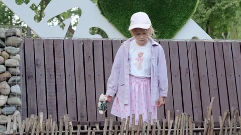 Young girl walking along rustic twig fence holding fabric doll in outdoor park Stock Footage 345361215
