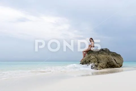 A young girl in a white swimsuit sits on a lonely stone on the beach. The wav Stock Photos