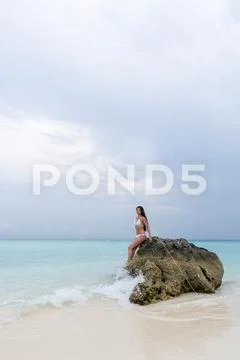 A young girl in a white swimsuit sits on a lonely stone on the beach. The wav Stock Photos
