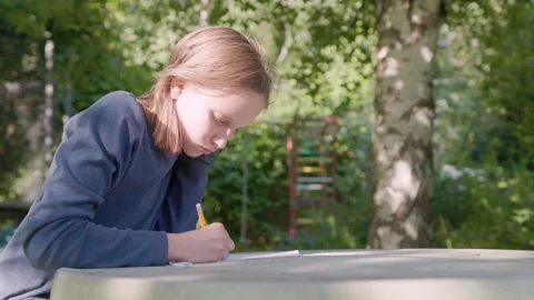 Young girl writes on notepad outdoors at table, showing signs of learning Stock Footage 346724693