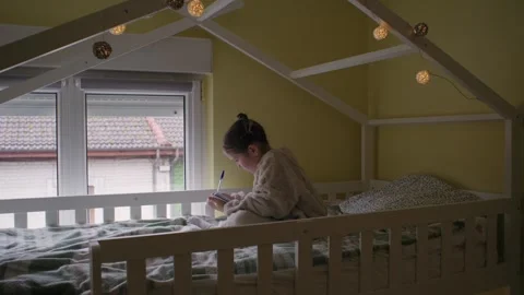 Young Girl Writing in a Notebook While Sitting on Her Bed Видео 333238824